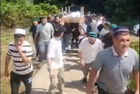 رجال يسيرون في موكب جنازة في منطقة خضراء، يحملون نعشاً على الأكتاف، مع جبال في الخلفية. يرتدون ملابس بسيطة وقبعات تقليدية، ويظهرون في حالة من الحزن والاحترام.