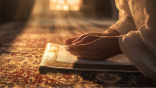 Person in traditional attire praying on a prayer rug with hands resting on an open Quran, illuminated by soft sunlight. The scene conveys spirituality and reverence.