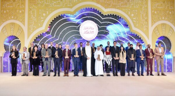 مشاركة مجموعة من الأفراد في حفل توزيع الجوائز، يقفون معًا على منصة مزينة بخلفية ملونة. يحمل كل شخص جائزة أو درعًا، ويظهرون بملابس رسمية. التصميم الخلفي يتضمن عناصر فنية ونصوص عربية.