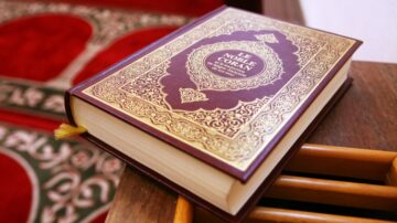 كتاب القرآن الكريم مفتوح على طاولة خشبية، مع غلاف مزخرف بالذهب. بالخلفية، سجادة حمراء مزخرفة.