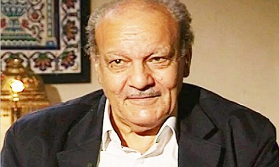 رجل مسن ذو لحية خفيفة يبتسم، يرتدي سترة داكنة وقميص أبيض، يجلس في بيئة داخلية دافئة مع خلفية مزخرفة.