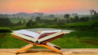 كتاب مفتوح على منصة خشبية مع منظر طبيعي خلفي يشمل تلال خضراء وسماء ملونة عند الغروب.