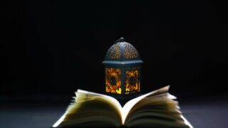 A decorative lantern with intricate patterns emits a warm glow beside an open book on a dark background, creating a cozy reading atmosphere.