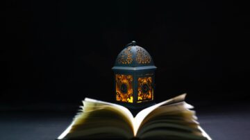 A decorative lantern with intricate patterns emits a warm glow beside an open book on a dark background, creating a cozy reading atmosphere.