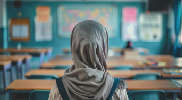 طالبة ترتدي حجابًا، تنظر إلى الفصول الدراسية الفارغة. الخلفية تحتوي على جدران زرقاء وأثاث مدرسي، مما يعكس بيئة تعليمية هادئة.