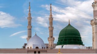 مسجد النبوي في المدينة المنورة، يظهر القبة الخضراء والمآذن الشاهقة تحت سماء زرقاء مع سحب خفيفة. تصميم معماري إسلامي يجسد الجمال الروحي والتاريخي للمكان.