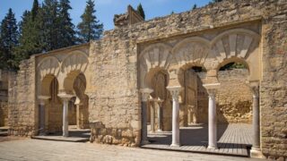 Historical ruins featuring stone arches and columns, set against a backdrop of trees and blue sky. The architecture reflects a blend of ancient design elements, emphasizing the texture of the weathered stone and wooden flooring.