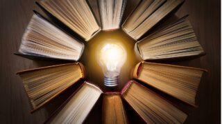 مصدر إلهام مع كتاب مفتوح ومصباح مضيء في المركز، محاط بكتب مغلقة مرتبة بشكل دائري. يعكس الصورة فكرة المعرفة والإبداع.