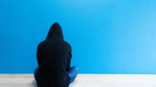 شخص يرتدي سترة سوداء وقبعة، يجلس على الأرض مواجهًا جدارًا أزرق، يعكس إحساسًا بالعزلة أو التفكير العميق.