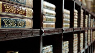 Close-up view of ornate, leather-bound books on wooden shelves, showcasing intricate gold lettering and decorative spines, highlighting the beauty of traditional Arabic calligraphy in a library setting.