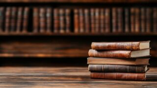 كتب قديمة مرتبة بشكل أنيق على سطح خشبي، مع خلفية تحتوي على مجموعة من الكتب الكلاسيكية في رفوف. تعكس الصورة جواً من الثقافة والمعرفة.