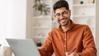 رجل شاب يرتدي نظارات وقميص بني، يجلس أمام جهاز كمبيوتر محمول ويبتسم أثناء الكتابة. تظهر خلفه رفوف تحتوي على نباتات وأغراض مكتبية، مما يضفي جوًا مريحًا ومناسبًا للعمل.