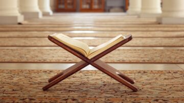 قرآن مفتوح على حامل خشبي في مسجد، مع خلفية تضم سجادات تقليدية وأعمدة معمارية، تعكس الأجواء الروحية والهدوء.