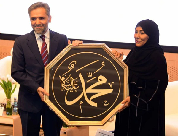 رجل وامرأة يحملان لوحة فنية تحمل اسم محمد عليه السلام بخط عربي، خلال حدث ثقافي. يبتسم كلاهما ويبدو في الخلفية جمهور ومكان مؤتمرات.
