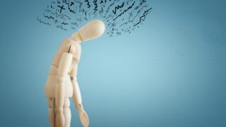 تمثال خشبي مائل للأمام، يعبر عن الإحباط أو القلق، مع رسومات متداخلة فوق رأسه تعكس مشاعر التوتر والضغط النفسي. الخلفية زرقاء بسيطة، مما يسلط الضوء على تعبير التمثال.