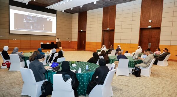 اجتماع عمل في قاعة مؤتمرات، حيث يجلس المشاركون حول طاولات مستديرة. متحدث يقدم عرضًا على شاشة كبيرة، مع وجود جمهور متنوع. بعض الحضور يستخدمون أجهزة الكمبيوتر المحمولة. الأجواء مهنية وتفاعلية.