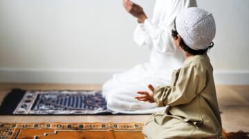 A child in traditional attire sits on a prayer mat, watching an adult praying. The adult is dressed in white, with hands raised in supplication, creating a serene atmosphere of faith and devotion. The setting features simple, neutral walls and wooden flooring, emphasizing the spiritual moment.