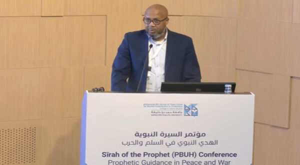 رجل يتحدث في مؤتمر السيرة النبوية، يركز على الإرشادات النبوية في السلام والحرب، مع وجود شاشة عرض أمامه.