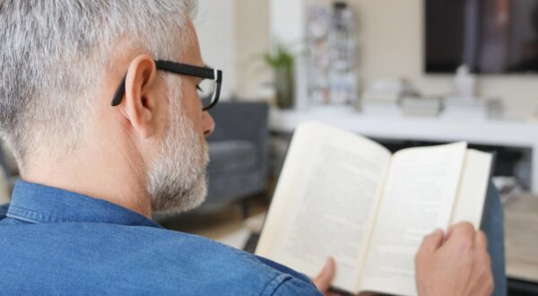 رجل مسن يرتدي نظارات يقرأ كتابًا في غرفة معيشة مريحة. تظهر خلفه أريكة رمادية وتلفاز. التركيز على الاستمتاع بالقراءة في أجواء هادئة.