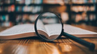 كتاب مفتوح مع عدسة مكبرة تركز على النص، خلفية مكتبة مليئة بالكتب.