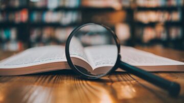 كتاب مفتوح مع عدسة مكبرة تركز على النص، خلفية مكتبة مليئة بالكتب.