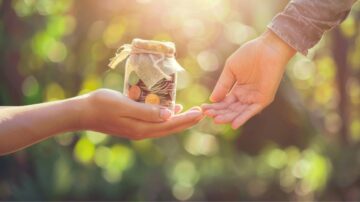 يد تُقدّم وعاء زجاجي مليء بالنقود المعدنية لشخص آخر، مع خلفية ضبابية من الطبيعة. يظهر مشهد تبادل العطاء والمشاركة بين اليدين في ضوء الشمس الدافئ.
