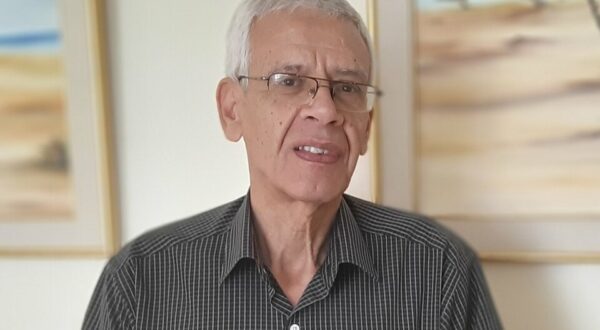 رجل مسن يرتدي قميصًا مخططًا باللونين الأسود والأبيض، يقف أمام جدار مزين بلوحات فنية تعكس مناظر طبيعية صحراوية. ملامح وجهه تدل على الحكمة والخبرة، ويظهر مبتسمًا.