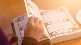 يد تمسك بكتاب مفتوح، يظهر نصوص باللغة العربية، مع صفحات تحتوي على آيات. الإضاءة الخلفية تضيف جواً دافئاً، مما يعكس لحظة من التأمل أو الدراسة.