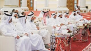 مجموعة من الرجال يرتدون الزي التقليدي في قاعة اجتماعات فاخرة، يجلسون على أرائك بيضاء. يتواجدون في بيئة رسمية مع زينة من الأزهار والطاولات اللامعة.