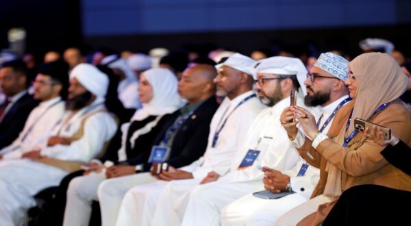 مشاركون في مؤتمر يرتدون ملابس تقليدية، يجلسون في صفوف، ويمتلكون بطاقات تعريف. يلتقط بعضهم الصور بهواتفهم، بينما يستمع الآخرون إلى المتحدث. الأجواء تعكس تنوع الثقافات في المناسبة.
