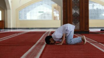 طفل صغير يؤدي سجود الصلاة في مسجد، يرتدي ملابس بسيطة، مع وجود سجادة حمراء في الخلفية ونوافذ مضاءة.