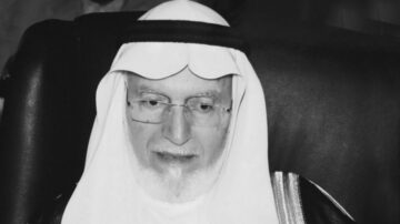 رجل مسن يرتدي الزي التقليدي العربي، يشمل الغترة والعباءة، يجلس مبتسمًا. تظهر ملامح وجهه بوضوح، مع نظارات وقصة شعر بيضاء. الخلفية غير واضحة وتحتوي على ظلال لأشخاص آخرين.