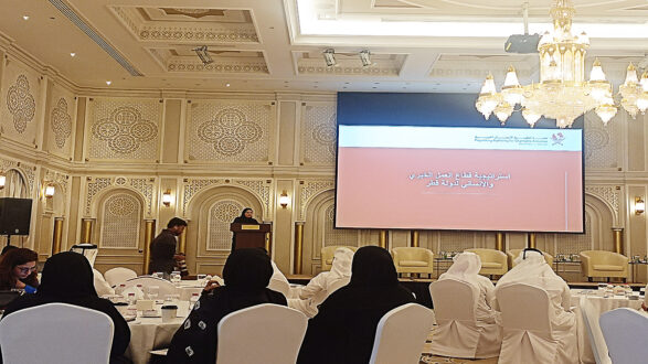 نسخة من مؤتمر أو ورشة عمل في قاعة أنيقة، حيث تتحدث امرأة على المنصة أمام جمهور يجلس حول طاولات. الخلفية تحتوي على شاشة عرض كبيرة تعرض عنوان الفعالية. القاعة مزينة بتفاصيل معمارية فاخرة وثريات مضيئة.