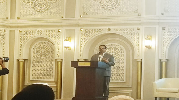 رجل يتحدث من منصة في مؤتمر داخل قاعة مزخرفة، مع تفاصيل معمارية فاخرة وإضاءة دافئة.