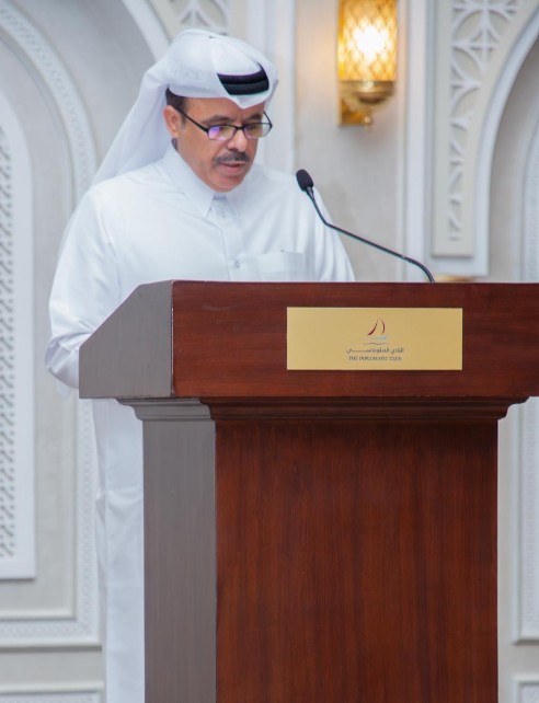 رجل في زي تقليدي قطري يقف خلف منصة ويتحدث إلى ميكروفون في حدث رسمي، مع خلفية مزخرفة وإضاءة دافئة.