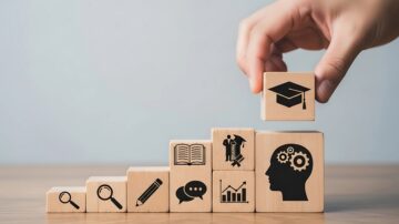 صورة توضح يدًا تضع مكعبًا خشبيًا مزخرفًا برمز قبعة التخرج على مجموعة من المكعبات المتزايدة. تمثل الرموز على المكعبات موضوعات مثل البحث، التعليم، والتفكير التحليلي، مما يعكس مفهوم التعلم والنمو الشخصي.