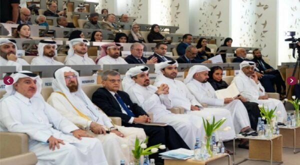 مجموعة من الرجال والنساء يرتدون ملابس تقليدية حديثة يجلسون في قاعة مؤتمرات، مع وجود لافتات تشير إلى مقاعد محجوزة. يركز الحضور على المتحدث في المنصة، مع وجود زهور في المقدمة. الأجواء تعكس حدثًا رسميًا يجمع شخصيات بارزة في المجتمع.