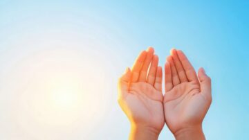 يدين مرفوعتين نحو السماء، مع خلفية زرقاء وصفراء تعكس ضوء الشمس. تعبر الصورة عن الأمل والإيمان.