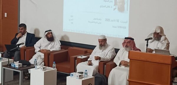 ندوة علمية تضم خمسة متحدثين، يجلسون على منصة، مع وجود شاشة خلفهم تعرض معلومات. المتحدثون يرتدون الملابس التقليدية، ويبدو عليهم التركيز أثناء النقاش. المقاعد مصنوعة من الخشب، وهناك زجاجات مياه على الطاولة.