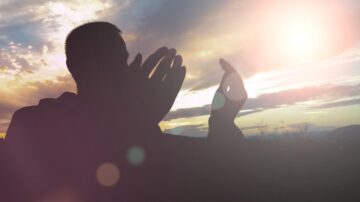 شخص يُظهر يديه في وضع تعبير تحت سماء مضيئة بألوان الغروب، مع أشعة الشمس تتسلل من بين الغيوم. الخلفية تحتوي على مناظر طبيعية هادئة، مما يخلق جوًا من التأمل والسكينة.