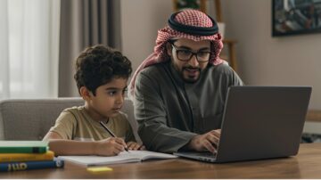 أب وابنه يجلسان على طاولة مع حاسوب محمول، الأب يساعد الابن في الدراسة. الغرفة تحتوي على نوافذ كبيرة وأثاث بسيط، مما يخلق جوًا مريحًا.