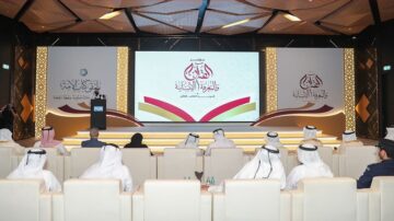 مؤتمر في قاعة حديثة تعرض شاشات كبيرة مع محتوى مكتوب باللغة العربية. الحضور يرتدون الملابس التقليدية ويجلسون في صفوف في المقدمة، بينما يتحدث متحدث على المنصة. تصميم داخلي أنيق مع إضاءة دافئة.