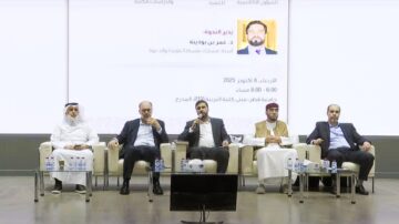 ندوة أكاديمية تُعقد في جامعة حمد بن خليفة، تضم خمسة متحدثين، بينهم رجل يرتدي بدلة سوداء يتحدث إلى الجمهور. يظهر على الشاشة خلفهم معلومات عن الندوة، بما في ذلك التاريخ 8 أكتوبر 2025، الساعة 6:00 مساءً.