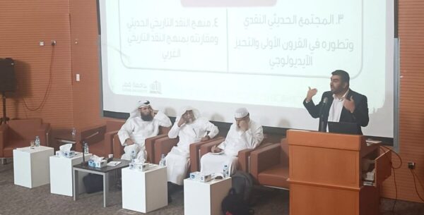 رجل يتحدث في ندوة بينما يجلس ثلاثة رجال آخرين في مقاعد. خلفية العرض تحتوي على نص عربي يتناول موضوع المجتمع الحديث وتطور القوانين. القاعة مجهزة بأجهزة عرض وأدوات للعرض.