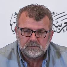 رجل متوسط العمر ذو لحية وشعر مجعد، يرتدي نظارات وقميص أزرق. يظهر بتعبير جاد في مؤتمر صحفي، مع خلفية تحمل نصوص باللغة العربية.