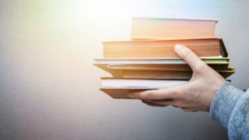 يد تحمل مجموعة من الكتب المكدسة، مع إضاءة خفيفة تبرز تفاصيل الكتب. الخلفية بسيطة وغير مزدحمة، مما يركز الانتباه على الكتب.