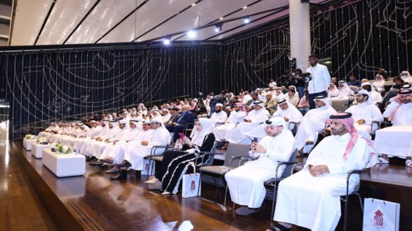 مجموعة من الحضور في قاعة مؤتمرات تتضمن رجالاً ونساءً يرتدون الزي التقليدي. يتواجد في المقدمة رجال يرتدون الغترة والعقال، بينما تجلس النساء في الخلف. القاعة مزينة بأضواء وخلفية فنية، مع وجود مصور في الجهة اليمنى.