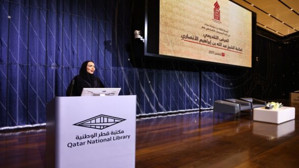 امرأة ترتدي عباءة سوداء تقف خلف منصة في مكتبة قطر الوطنية. خلفها شاشة تعرض نصًا باللغة العربية يتعلق بحدث ثقافي. الإضاءة داخل القاعة دافئة والأرضية خشبية.