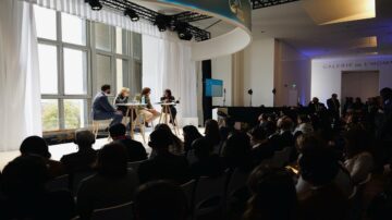 جلسة نقاشية في حدث مع متحدثين يجلسون على منصة أمام جمهور كبير. يوجد إضاءة طبيعية من النوافذ الكبيرة.