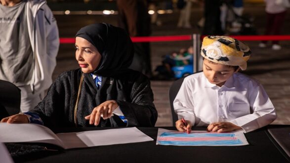 امرأة ترتدي عباءة وحجاب تجلس بجانب طفل صغير يرتدي ثوباً وعقالاً، حيث يقوم الطفل برسم على ورقة بيضاء. الخلفية تحتوي على أضواء خافتة وأشخاص آخرين في المكان.